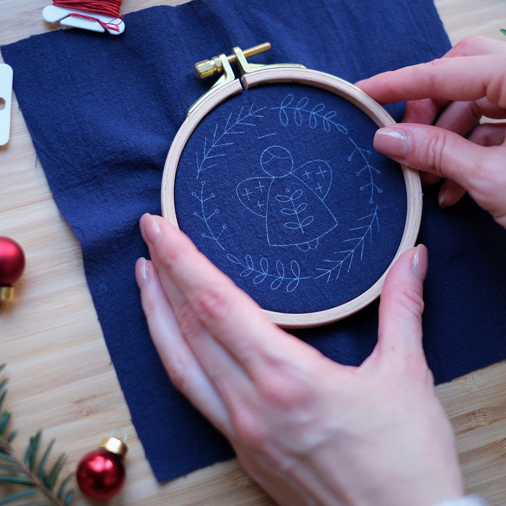 Embroidery hoop with a design on navy fabric, surrounded by Christmas decorations on a wooden surface.